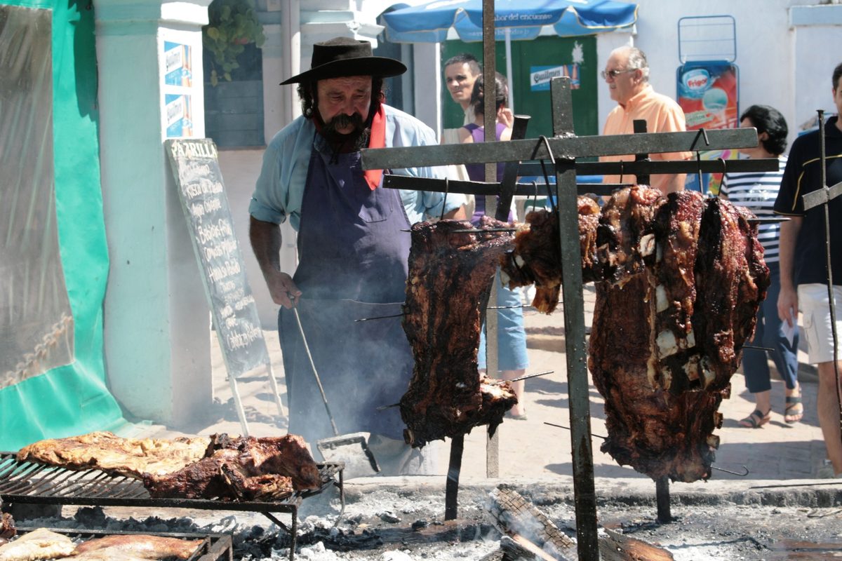 Gaucho in a black hat grilling an asado over open coals at a festival in San Antonio de Areco