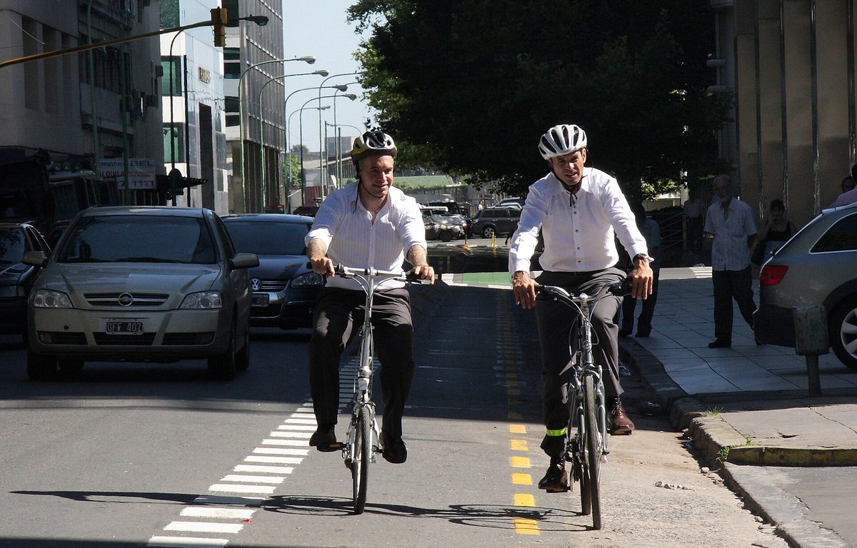 Two helmeted cyclists riding in a protected ciclovia bike lane beside car traffic on a Buenos Aires street
