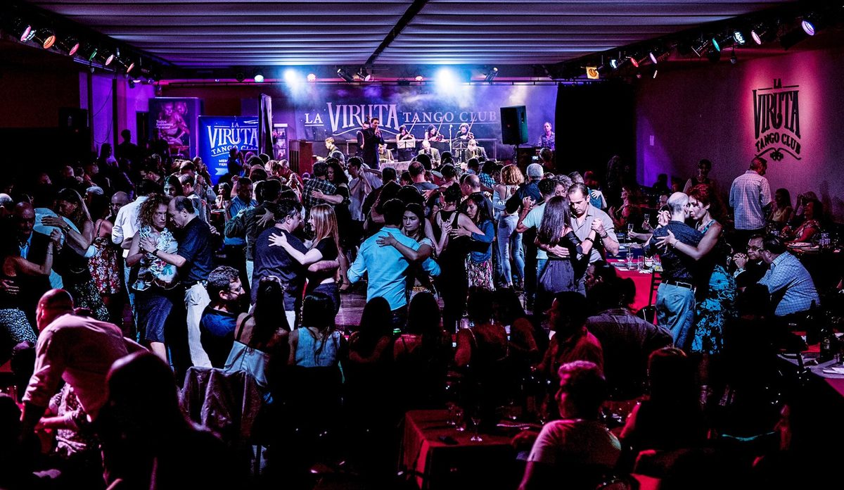 A packed social milonga floor in Buenos Aires with couples in close embrace and tables around the edge